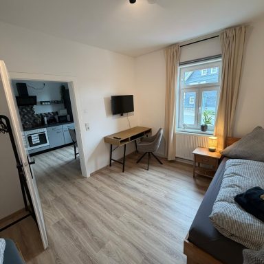 Kleines Zimmer mit Bett, Schreibtisch und Küchenbereich im Hintergrund. Helle Wohnatmosphäre.