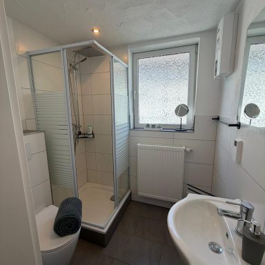 Kleines Badezimmer mit Dusche, Waschbecken und Toilette, helles Fenster sorgt für Licht.