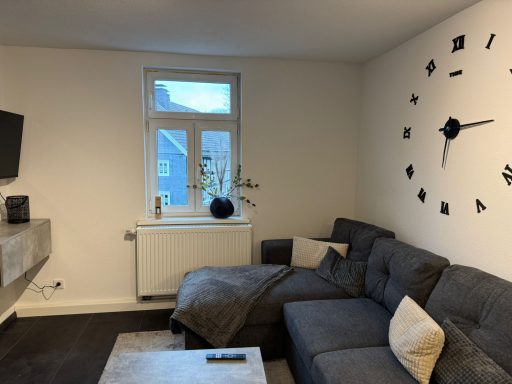 Moderne Wohnzimmer mit grauem Sofa, großer Uhr und Fenster mit Aussicht.